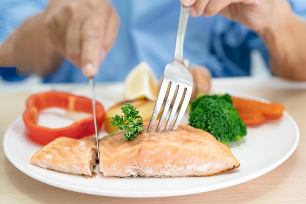 Asian senior woman patient eating Salmon steak breakfast with vegetable healthy food while sitting and hungry on bed in hospital