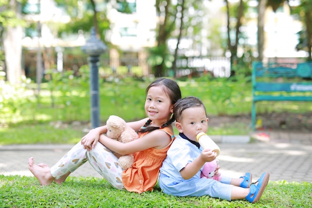 Asian sister and younger brother in the garden. Child girl cuddle teddy bear doll and boy Suck milk from bottle.