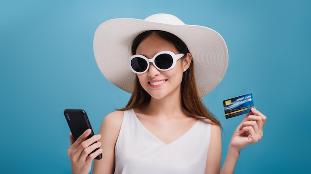 Asian smiling beautiful women wear a wide hat and sunglasses using smartphone shopping online with credit card isolated in blue color backgroundConcept of Travel business with Promotion and Sale