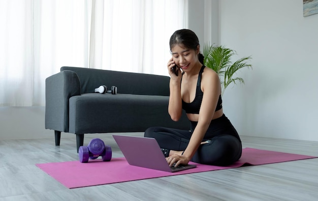 Asian sporty woman in sportswear working out and using laptop and call phone at home in living room sitting on the floor with dumbbells on yoga mat Sport and online training concept