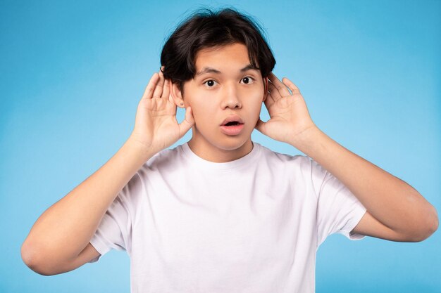 Asian teen guy listening holding hands near ears blue background