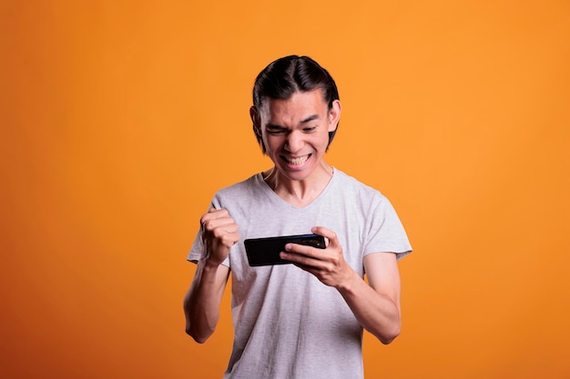 Asian teenager winning video games on phone, showing victory gesture, happy young man enjoying online mobile videogames. Gamer playing, using smartphone entertainment application