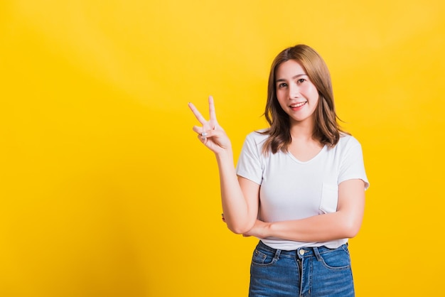 Asian Thai happy portrait beautiful cute young woman smile standing wear t-shirt showing finger making v-sign symbol near eye looking to camera, isolated studio shot yellow background with copy space