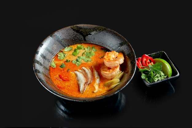 Asian tom yum soup with shrimps, shiitake mushrooms, octopus, tomatoes and rice in a plate. Classic Asian cuisine. Food delivery. Isolated on black