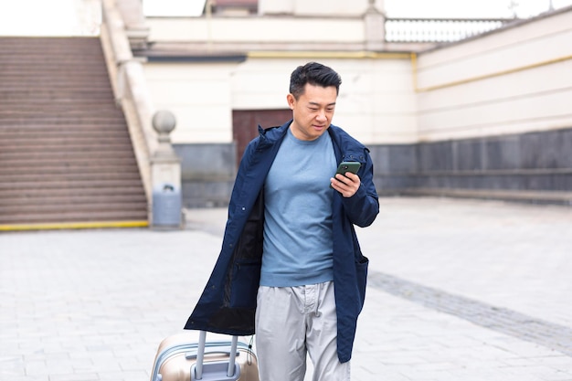 Asian tourist male passenger arrives from the airport walks around the city with a suitcase a man smiles and rejoices uses the phone and the application to book accommodation and book a taxi