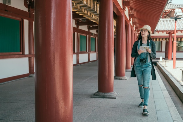 Asian tourist woman with map paper searching for attractions travel in osaka japan visit shitennoji. full length of young girl backpacker with camera walking in historical japanese red wood building