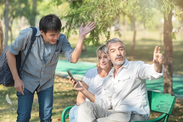 Asian tourists young man with smart mobile phone in his hands asks for directions from the old caucasian elder man who sit with woman, He pointed the finger to the way.