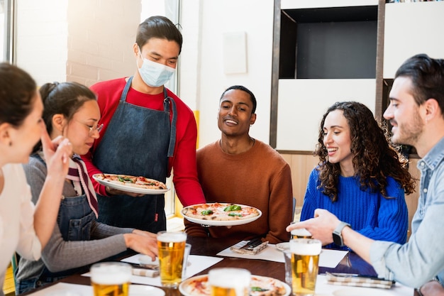 Asian waiter wearing protective face mask serving pizza to a multiracial group of young friends