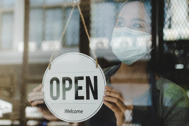 Asian waitress staff woman turning open sign board on glass door with bokeh light in cafe coffee shop for lift lockdown hotel service cafe restaurant small business owner food and drink concept