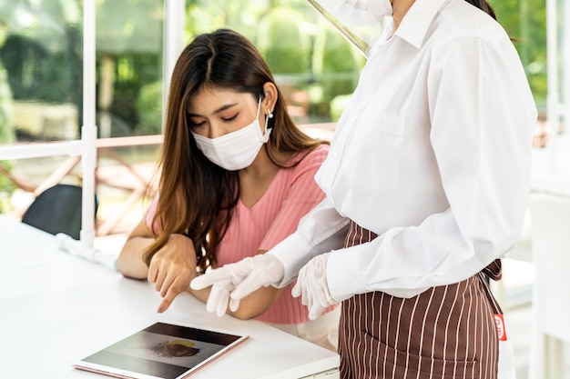 Asian waitress wear face mask and face shield using tablet to show restaurant electronic menu