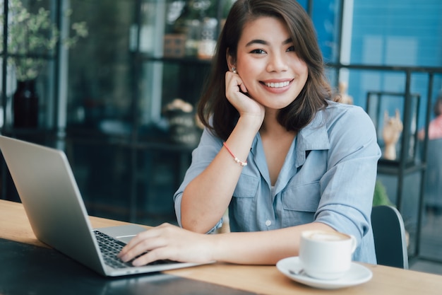 Asian woman in blue shirt  in cafe drinking coffee and using laptop computer working business online marketing