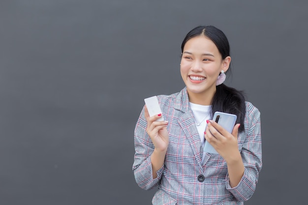 Asian woman in brown striped blazer smiles happily successful while holds smartphone credit card