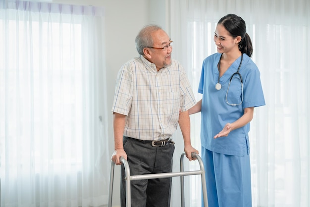 Asian woman caregiver doctor helping elderly man patient to walk using walker frame at home