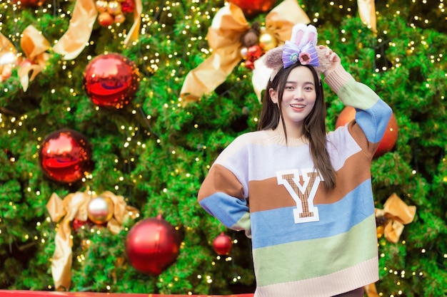 Asian woman in a colorful sweater is smiling happily in front of the Christmas tree In the theme.