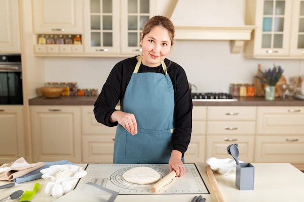 Asian woman cook in apron look at camera sprinkles flour on cooked wheat pastry dough on silicone baking mat in modern kitchen