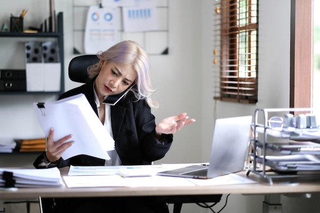 Asian woman cover her face with hand and feel upset while talk on mobile phone with customer in front of laptop computer on desk at officeStress office lifestyle concept