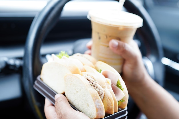 Asian woman driver hold ice coffee cup and sandwich bread for eat and drink in car dangerous and risk an accident