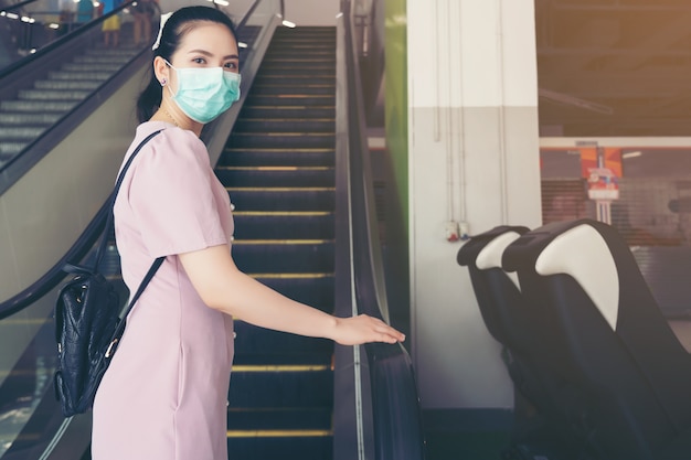 Asian Woman hand holds escalator handrail, coughs in her elbow and keep social distancing from other people with surgical face mask protection - she commutes in the shopping mall