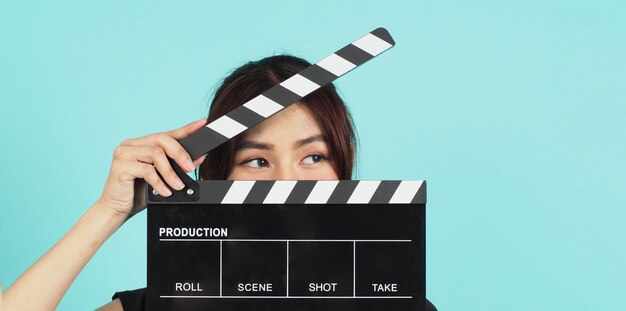 Asian Woman holding black clapperboard and closing face on mint or Tiffany Blue background.