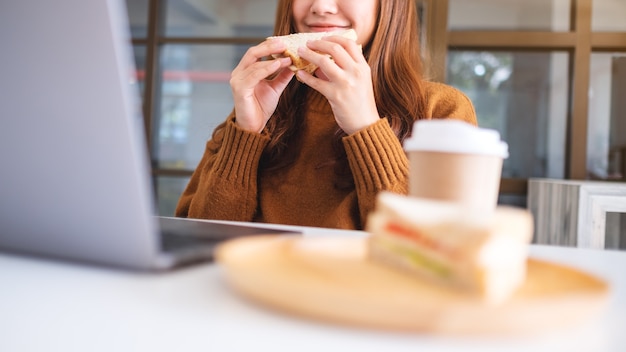 An asian woman holding and eating whole wheat sandwich while working on laptop computer