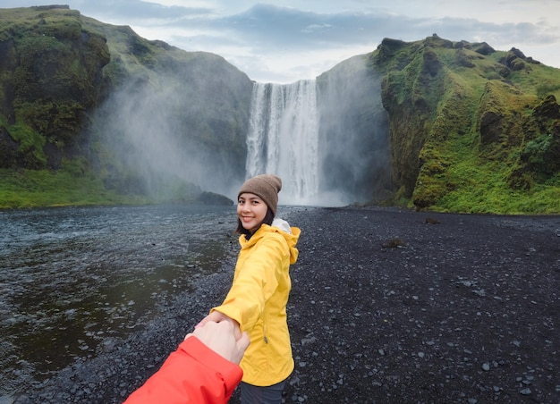 Asian woman holding hands with couple at front of Skogafoss waterfall flowing on cliff in summer at Iceland