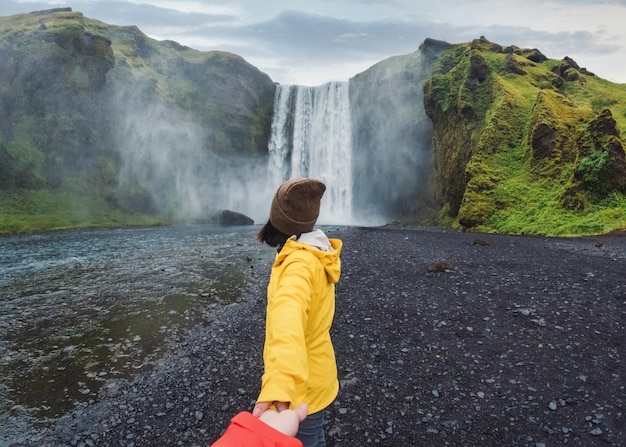 Asian woman holding hands with couple at front of Skogafoss waterfall flowing on cliff in summer at Iceland