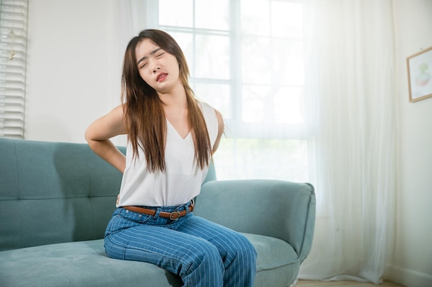 Asian woman holding her lower back feeling painful waist hurt massage in living room, beautiful young female touching back pain sitting in sofa she suffering from backache at home, healthcare medical