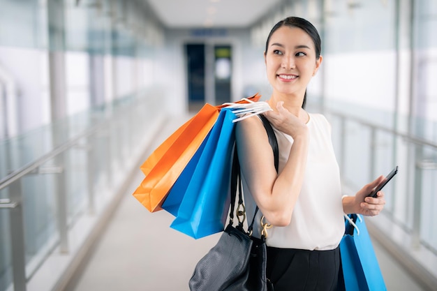 Asian woman holding smart phone with shopping bags walks through the city streets. Summer Style.