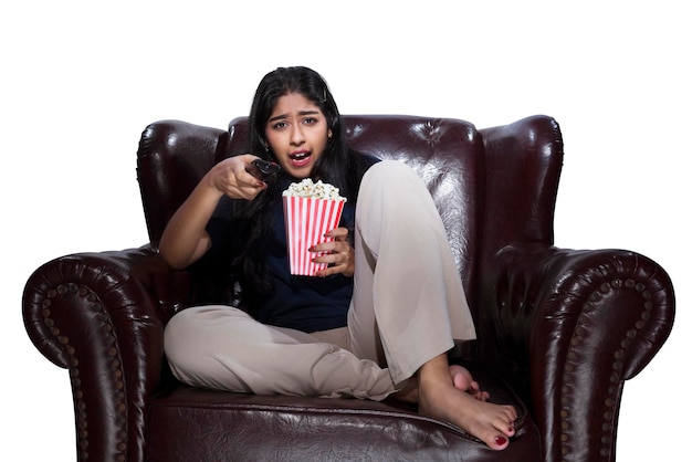 Asian woman holding a television remote and popcorn sitting on the couch with a scared expression