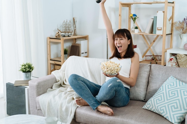 asian woman is sitting on the couch watching a competitive olympic game at home.  japanese girl with popcorn shouted loudly because her team just scored a point.