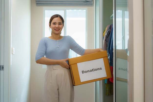 Asian woman is standing near closet of clothes in the dressing room carrying a box of clothes donated to take to the donation center.