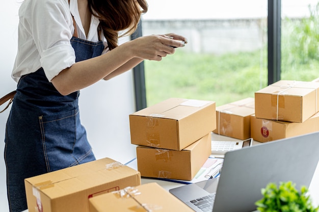 An Asian woman is using a smartphone to take pictures in front of a parcel box for evidence, she owns an online store, she packs and ships through a private shipping company. Online selling concept.