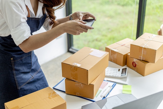 An Asian woman is using a smartphone to take pictures in front of a parcel box for evidence, she owns an online store, she packs and ships through a private shipping company. Online selling concept.