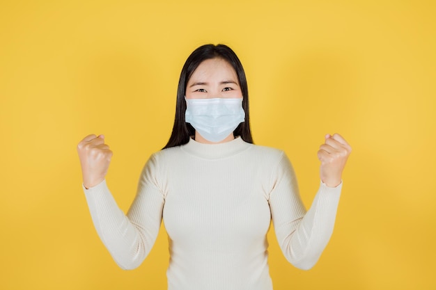 Asian woman in medical face mask to protect Covid19 Coronavirus raise hands glad excited cheerful on yellow background