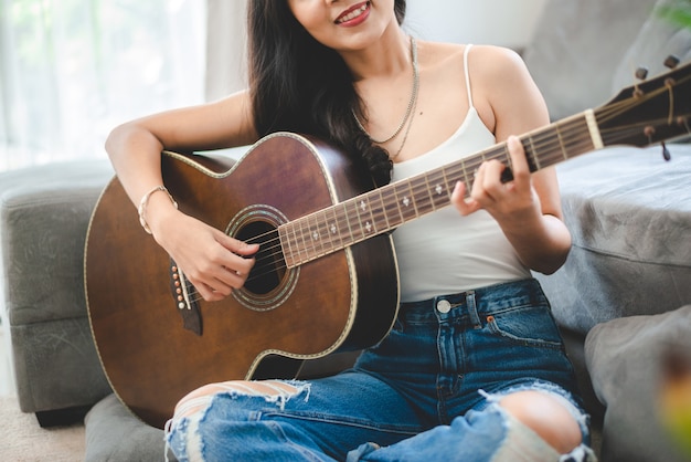 Asian woman playing music by guitar at home, young female guitarist musician lifestyle with acoustic art instrument sitting to play and sing a song making sound in hobby in the house room