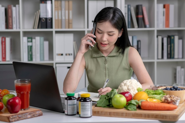 Asian woman professional nutritionist consulting with her client on the phone surrounded by a variety of fruits nuts vegetables and dietary supplements on the table