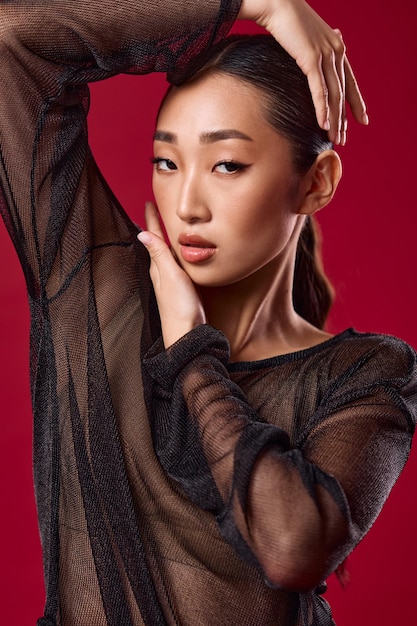 Photo asian woman in sheer top posing with hands on head against red background hair pulled back