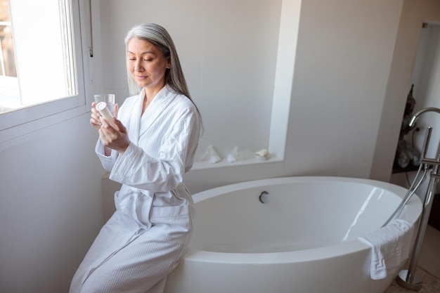 Asian woman sitting on bathtub in bathroom holding cosmetic jar