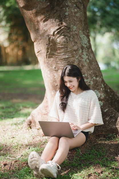 Asian woman sitting on picnic and lawn at park working on laptop Asian female using laptop while sitting under a tree at park with bright sunlight Work from anywhere concept