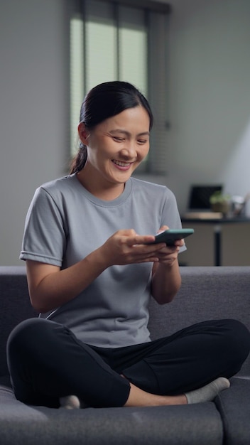 Asian woman sitting on sofa and holding smart phone at home Happy woman using smart phone for searching shopping online or surfing social media