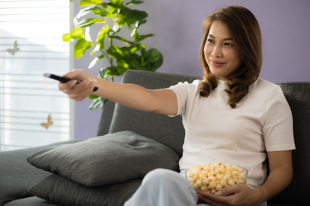 Asian woman sitting on sofa with relax and cozy gesture holding remote control aiming to tv, watching television and smile with fun and happy in living room with sunlight and light flare.