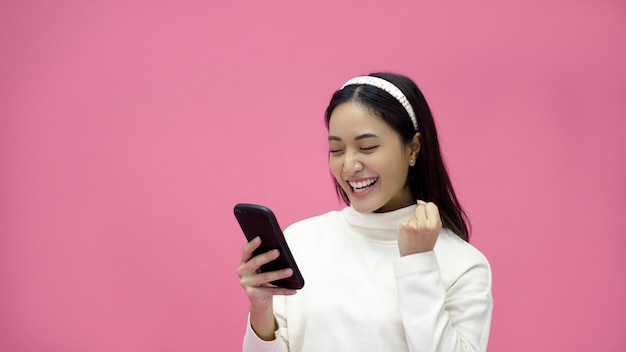 Asian woman smiling and using mobile phone with tablet and excited for online shopping on isolated pink background