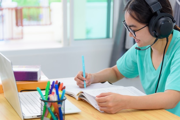 Asian woman student teenage girl with glasses headphones sitting happy smile looking writing notes at book laptop computer on table learning online study education from the class of university at home