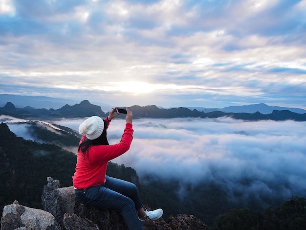 Asian woman tourist sitting on the rock at the top of mountain and taking photograph sunrise sky.