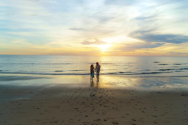 Asian woman traveler holding man's hand and looking beautiful sunset on the beach, Couple on vacation in summer and freedom concept