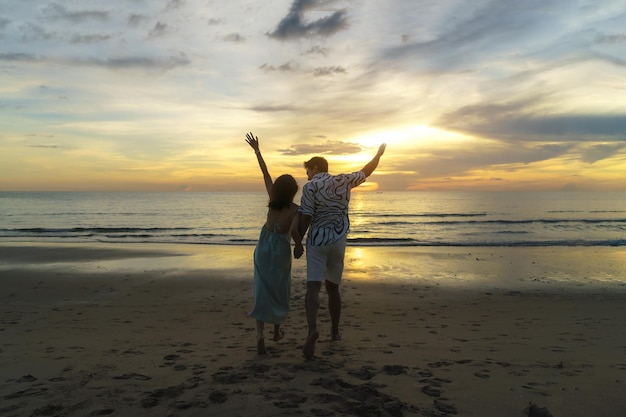 Asian woman traveler holding mans hand and looking beautiful sunset on the beach