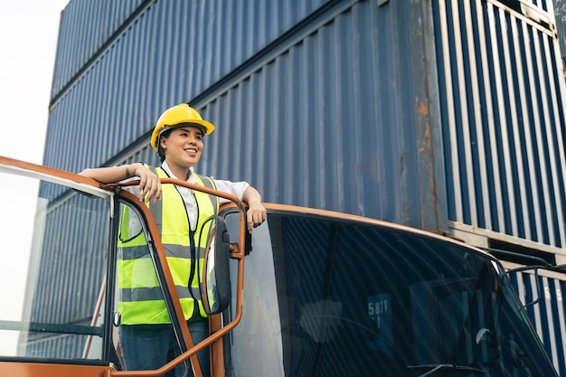 Asian woman truck driver wear yellow helmet and safety vest standing in front of truck vehicles.