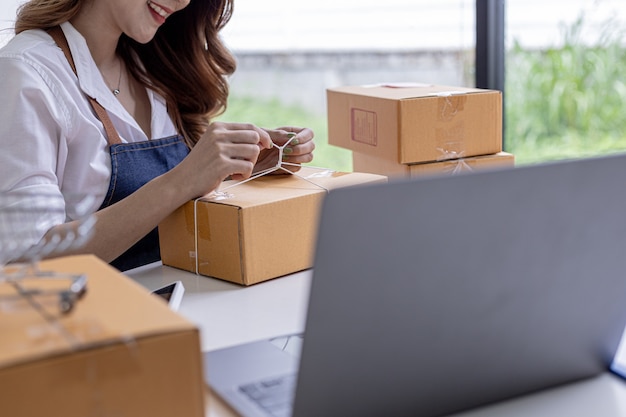 An Asian woman tying a parcel to a customer's box, she owns an online store, she packs and ships through a private transport company. Online selling and online shopping concepts.