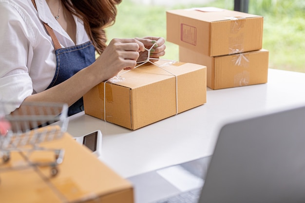 An Asian woman tying a parcel to a customer's box, she owns an online store, she packs and ships through a private transport company. Online selling and online shopping concepts.