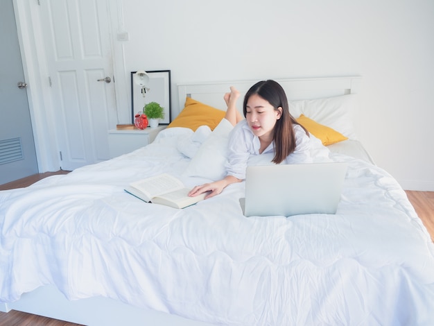 Asian woman using computer and read book on bed in the morning